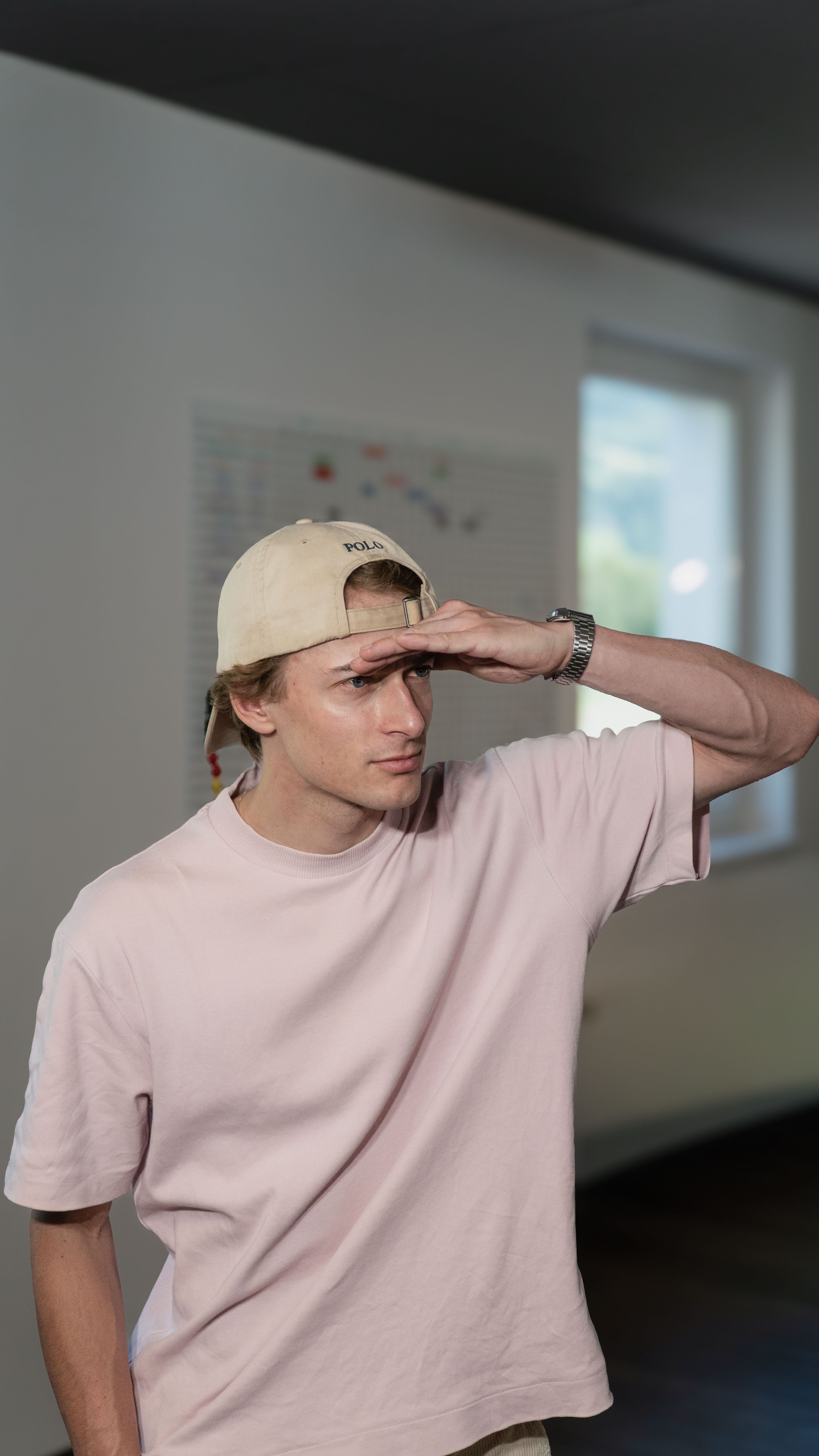 Ein junger Mann mit Kappe und rosa T-Shirt schirmt die Augen mit der Hand ab und blickt suchend zur Seite.