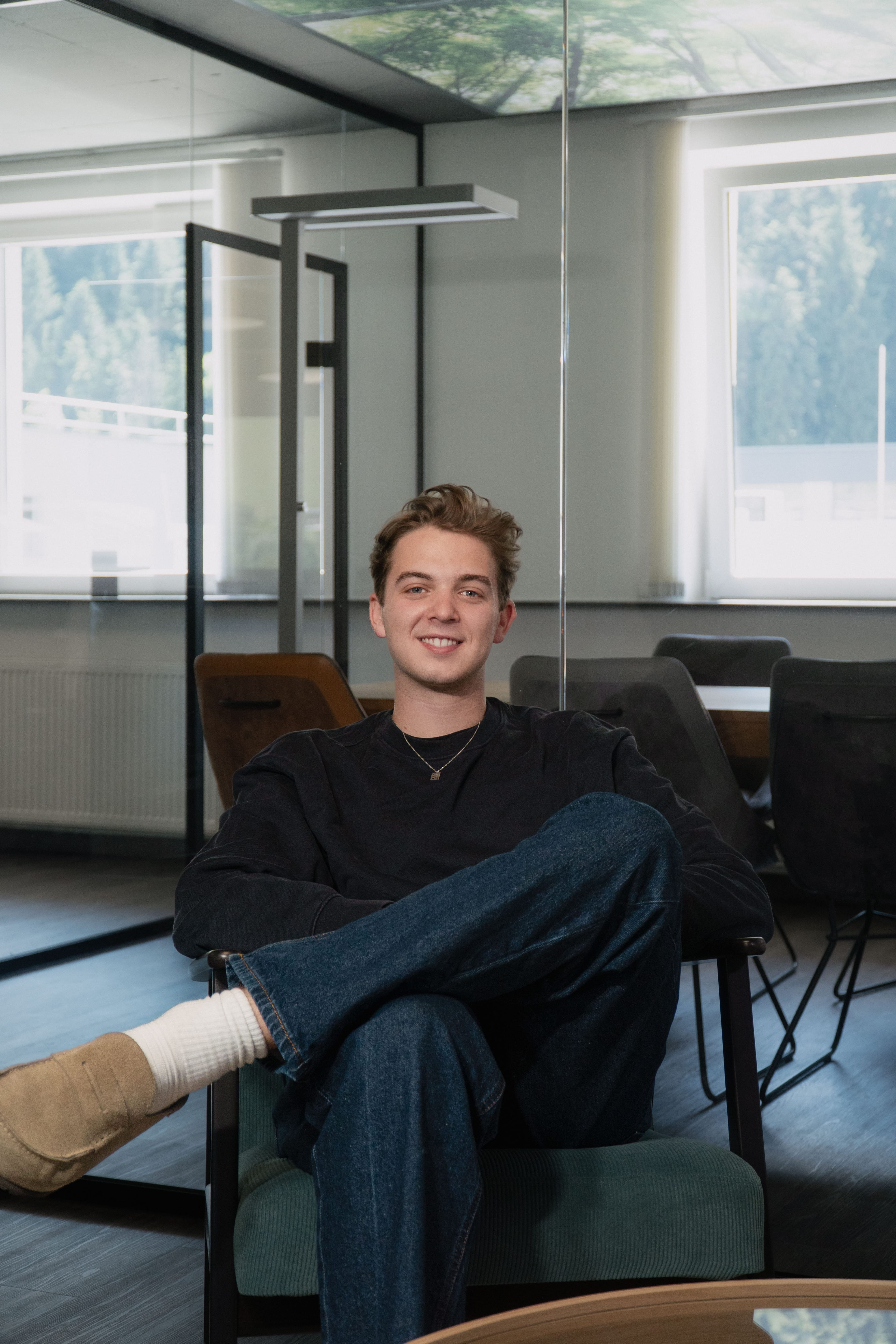 Ein lächelnder junger Mann sitzt mit überschlagenen Beinen in einem Sessel in einem modernen Büro.