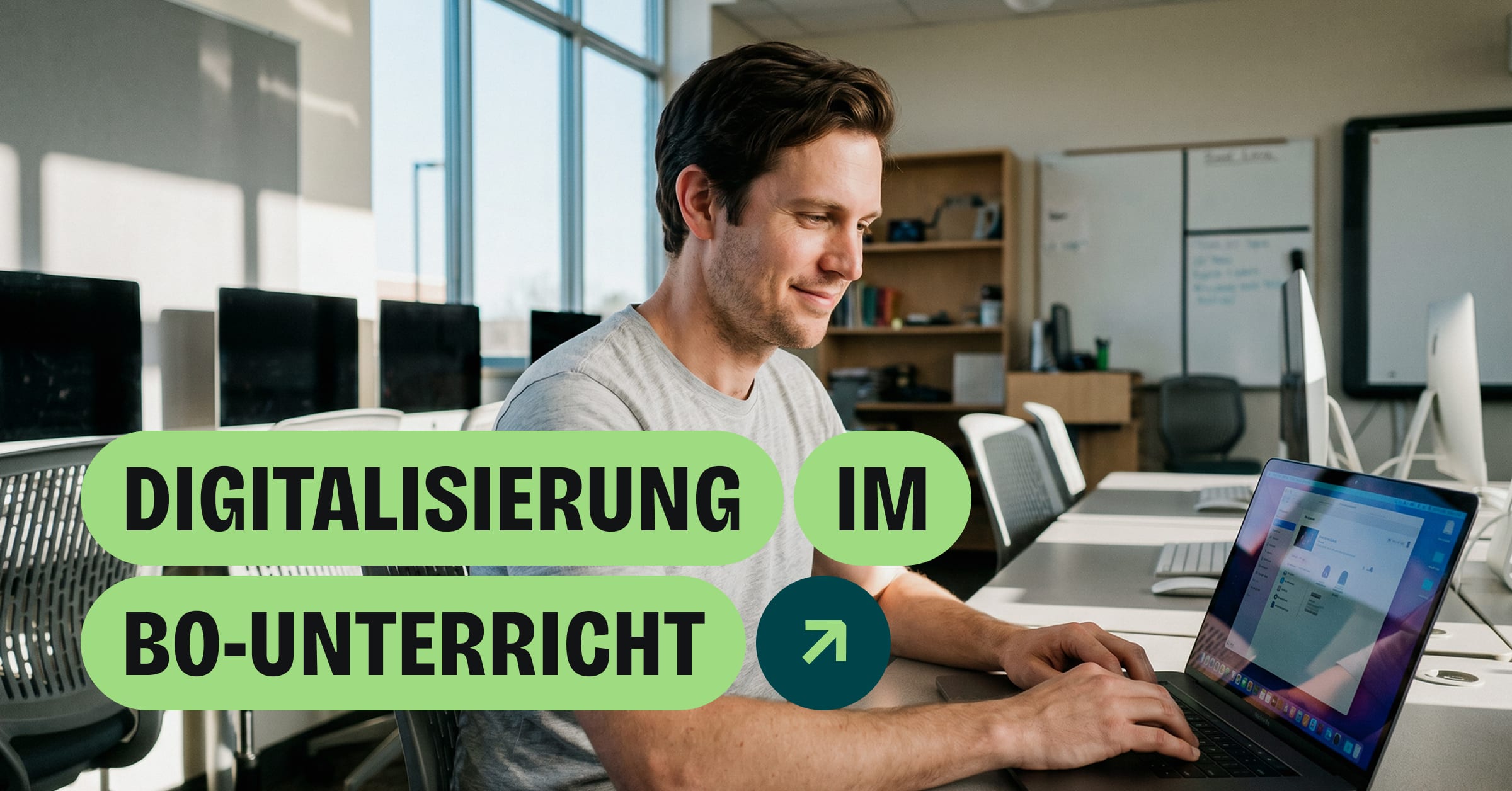 Ein lächelnder Mann arbeitet am Laptop in einem Computerraum mit mehreren PC-Arbeitsplätzen im Hintergrund.