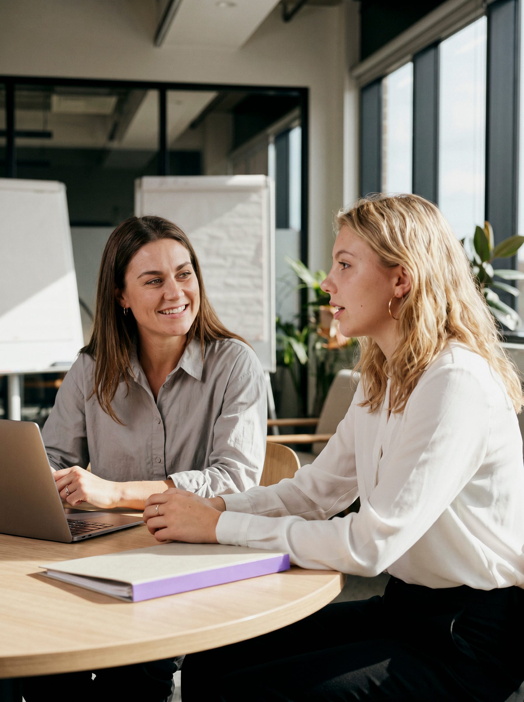 Zwei Frauen sitzen an einem runden Holztisch mit Laptop und Ordner und unterhalten sich im Büro.